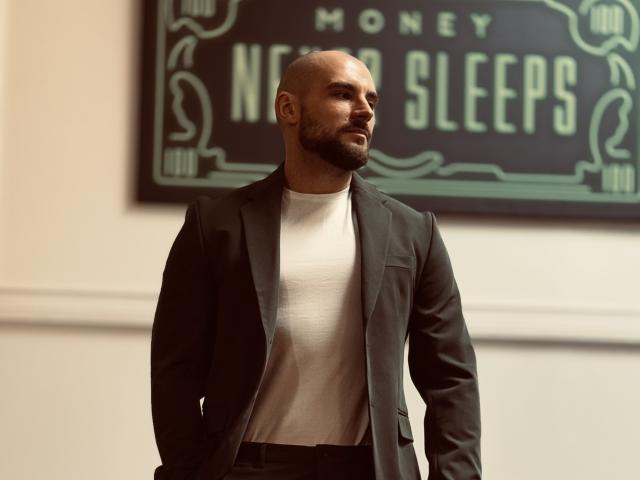 AdamJameson, bald with a beard, wearing a black suit over a white t-shirt, standing confidently with hands in pockets, looking to the side, in a well-lit room.