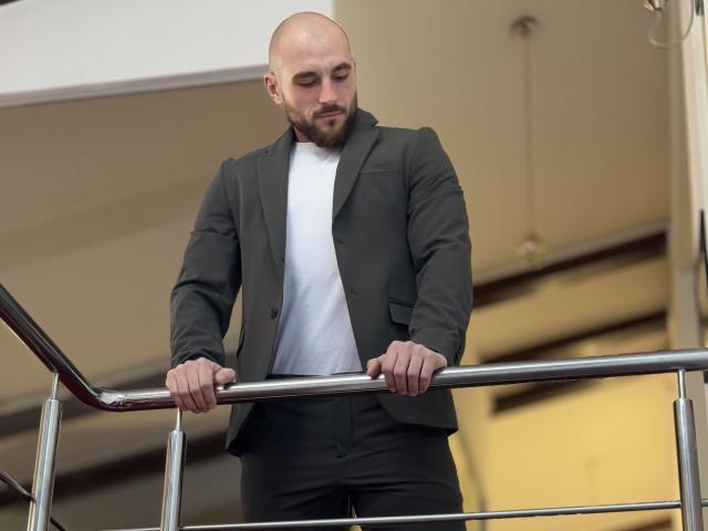 AdamJameson, bald with a beard, wearing a dark green suit and white shirt, leans on a metal railing with both hands, looking downward.