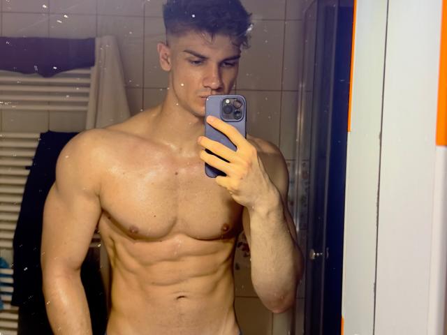 Nude MaxKaiden with a muscular, toned body, visible abs, and small nipples, holding a phone, taking a mirror selfie, showing a slight bulge, short curly brown hair.