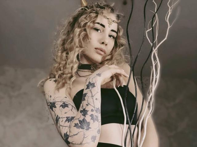 LunaBell with curly blonde hair, black strapless top, tattooed arm, choker, and headpiece, holding white and black sticks, looking seductive. Boobs, tattoos, and a cute face.