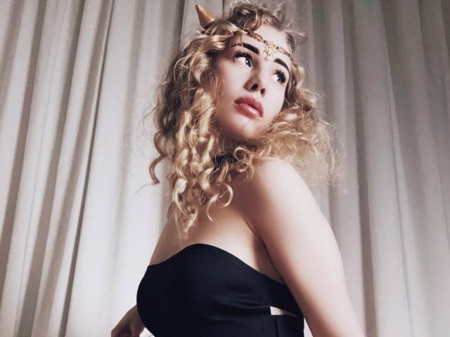 LunaBell with curly blonde hair, black strapless top, gold headpiece, and small horn, looking up with pouty lips, showing her tits slightly. Soft light, neutral background.