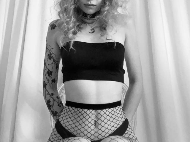 Black and white photo of LunaBell with curly hair, wearing a strapless top, fishnet pants, and a choker. She has tattoos on her left arm and a belly button piercing.