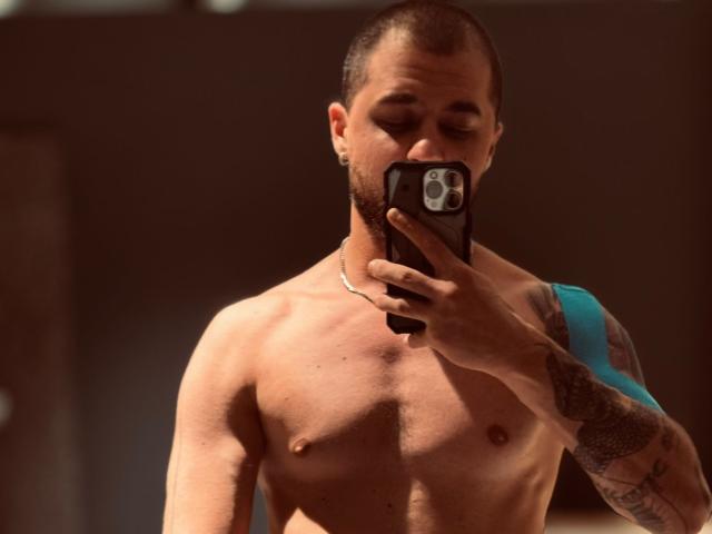 Shirtless JamesMyers with a trimmed beard, holding a phone in front of his face, tattoo on right arm, and a silver necklace, taking a mirror selfie.