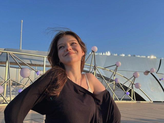 KarenaHoltgrewe with long brown hair, smiling, wearing a black off-shoulder top, showing her tits, standing on a wooden deck with pink and purple balloons in the background.