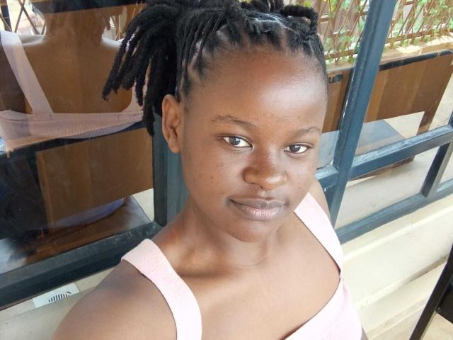 CuteBetty with dark brown skin, small nose, and small shiny lips, wearing a pink tank top, has medium-sized tits, and short, neat dreadlocks, standing near a window.