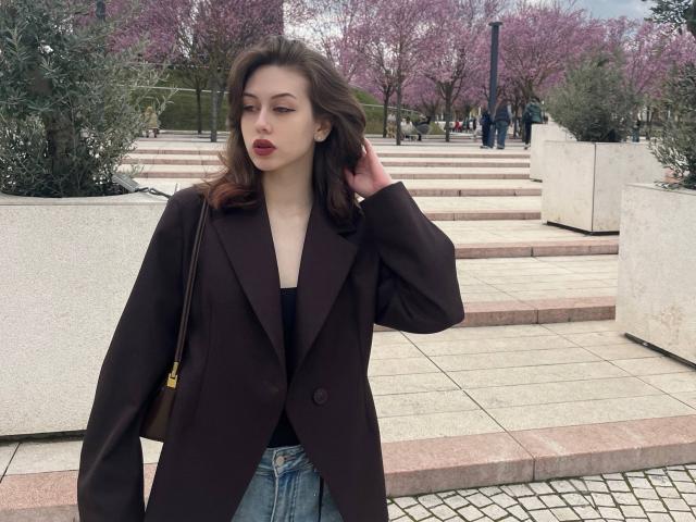 RussellMixer hat braunes Haar, roten Lippenstift, einen dunkelbraunen Mantel, ein schwarzes Oberteil, blaue Jeans und eine braune Tasche. Sie steht auf rosa und weißen Steinstufen vor lila Bäumen..