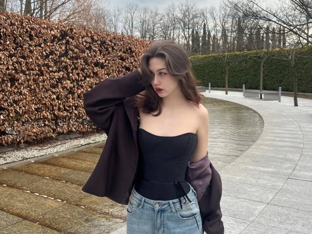 RussellMixer con cabello castaño ondulado, lápiz labial rojo, top negro sin tirantes, jeans de mezclilla y abrigo oscuro, de pie junto a un camino de piedra curvo, con la mano en el abrigo. Se ven los senos, los pechos son pequeños..
