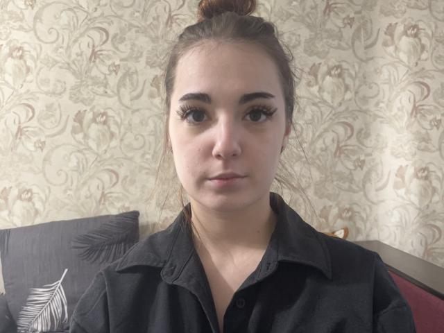 MarietteHot has long brown hair in a bun, wearing a black shirt, with thick black eyelashes and a neutral expression, showing her pouty ass-kissing lips.