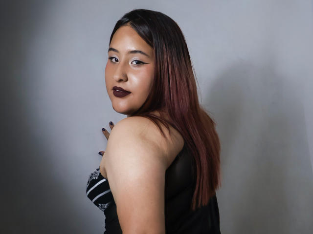 RavenPurpple with dark lipstick, black eyeliner, and straight brown hair, looking over shoulder, wearing a black strapless top, showing her bare shoulder and black-painted nails.