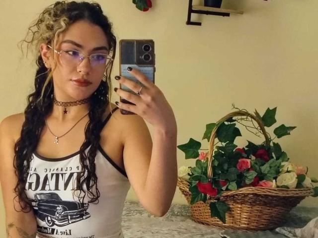 AdaraAngel with curly black and blonde hair, glasses, choker, tank top, taking a selfie, holding phone, wicker basket with roses and green leaves on the right.