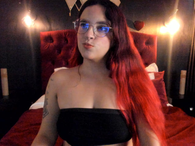 LaiaJonnes, avec de longs cheveux roux, des lunettes, un haut noir sans bretelles, un tatouage sur le bras droit, regardant la caméra, debout devant une tête de lit en velours rouge, un lit rouge, avec des lumières chaudes..