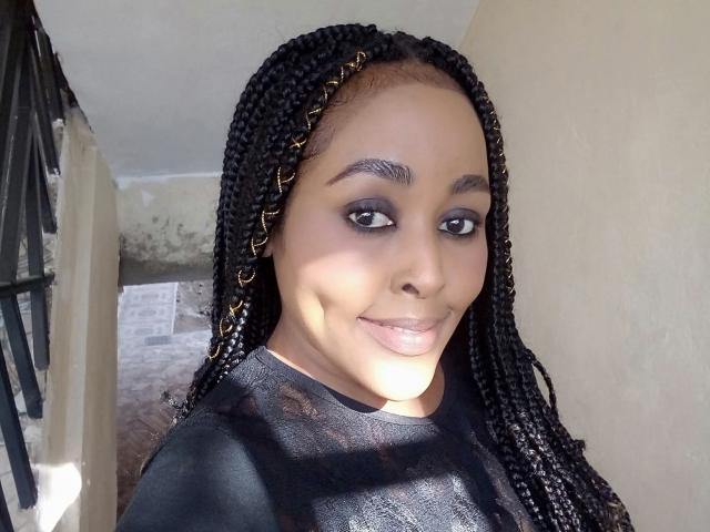SexyTiffa with long black braids, wearing a black top, smiling at the camera, has dark eye makeup, and a cute, confident face, standing in a sunlit room.