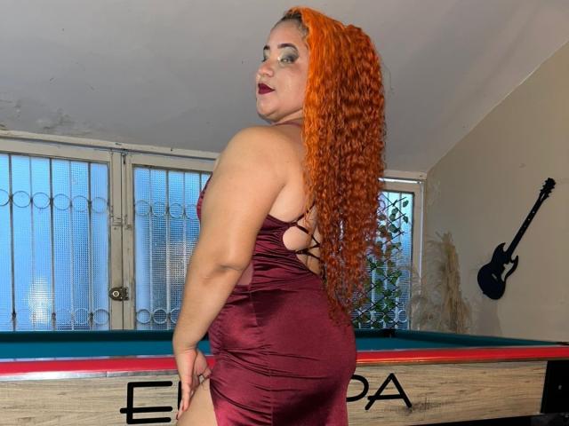SecretFroomies with long red curly hair, wearing a tight maroon dress, showing her ass, standing next to a pool table, looking back over her shoulder.