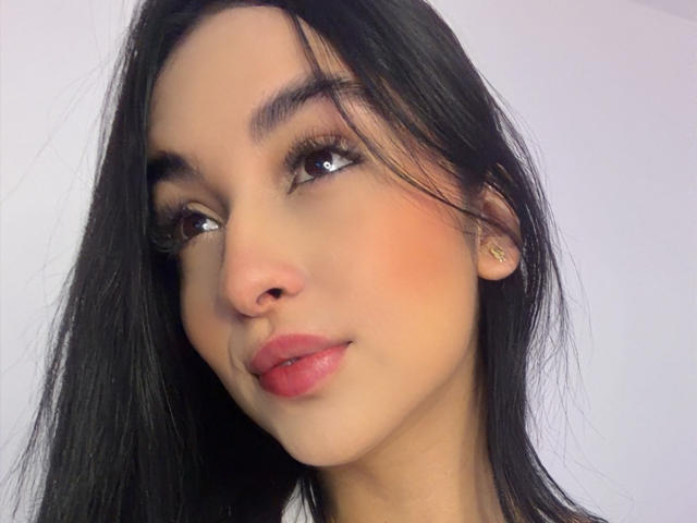 EmmaWaltter's face with long black hair, brown eyes, pink lips, subtle blush, small gold earrings, and a slight smile. Her nose is pierced. She looks to the side.