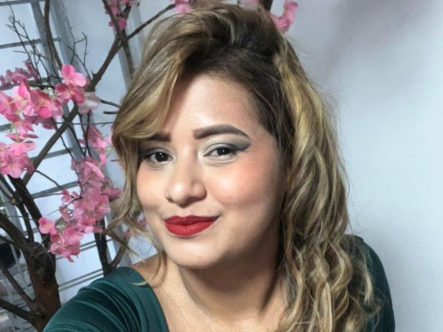 A photo of SecretFroomies with wavy blonde hair, red lipstick, and green top, smiling at the camera, with pink cherry blossoms in the background.