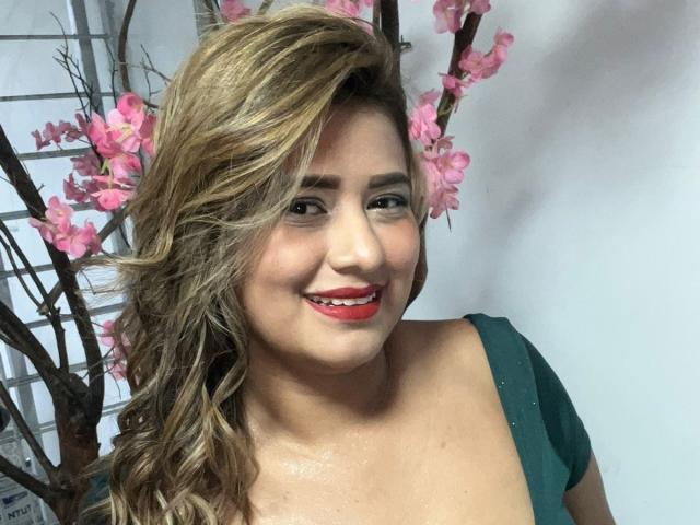 SecretFroomies with wavy blonde hair, red lipstick, and green dress, smiling at the camera, pink cherry blossoms in the background, showing off her shiny tits and cute ass.