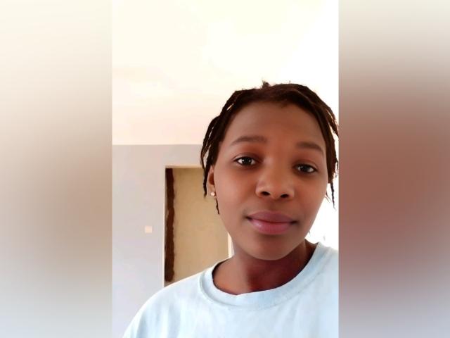 Rubiya with dark brown skin, small round nose, and short braided hair, wearing a white shirt, smiling softly at the camera, in a bright indoor setting.