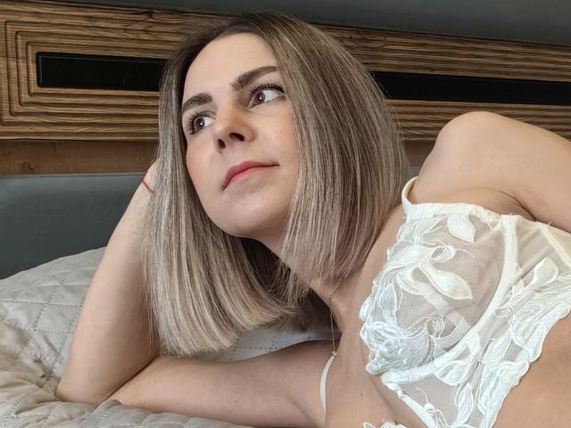 IceMaya with blonde bob, looking up, wearing white lace top, lying on bed, head on hand, soft smile, bed with white quilt, wooden headboard.