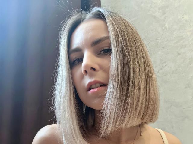 IceMaya with a short, straight blonde bob, light skin, and subtle makeup, looking slightly confused with parted lips and small nose, wearing a thin-strapped top.