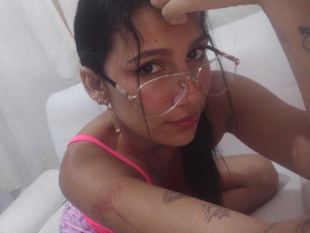 KataleyaMansson with glasses, nose ring, pink tank top, tattoos, and long dark hair, touching her forehead, showing off her tits, and a slight smile.