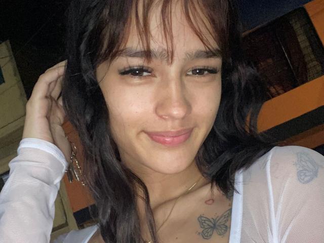 MelodyMansson with dark brown hair, slightly wavy, smiling softly. She's wearing a white, sheer top, showing small tits and a butterfly tattoo on her chest. Hands in her hair.