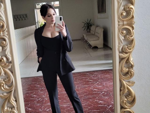 EmmaNoa, con cabello oscuro, un blazer negro y pantalones negros, está tomando una selfie frente a un espejo, mostrando sus grandes senos y con las manos en los bolsillos, de pie sobre un suelo cubierto de alfombra roja..