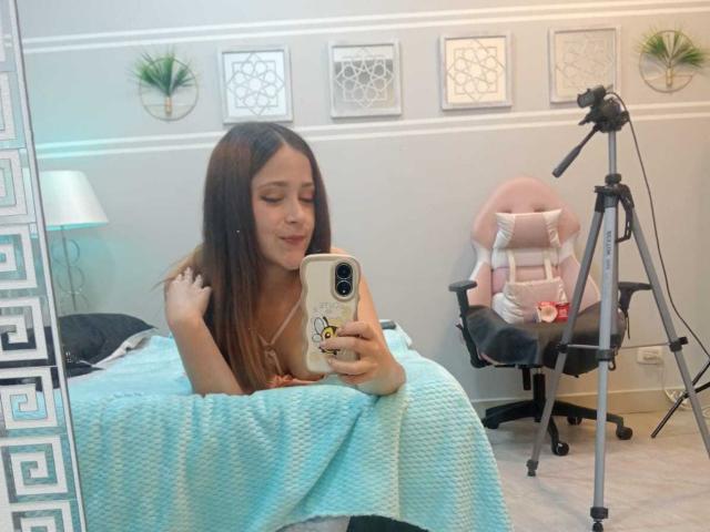 ElisabetLot with long brown hair, smiling, holding a phone, lying on a bed with a blue blanket, in front of a mirror, near a tripod and pink chair.