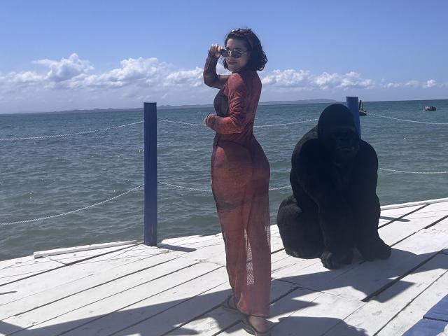 HannaBorbon è in piedi su un molo di legno, indossa un abito rosso trasparente a maniche lunghe, con il sedere e i seni visibili, e porta degli occhiali da sole. Alla sua destra si trova una statua nera..