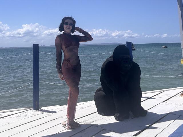 HannaBorbon in a sheer, red mesh bodysuit, standing with a black gorilla on a white wooden dock, wearing sunglasses, hand on head, blue ocean in background.