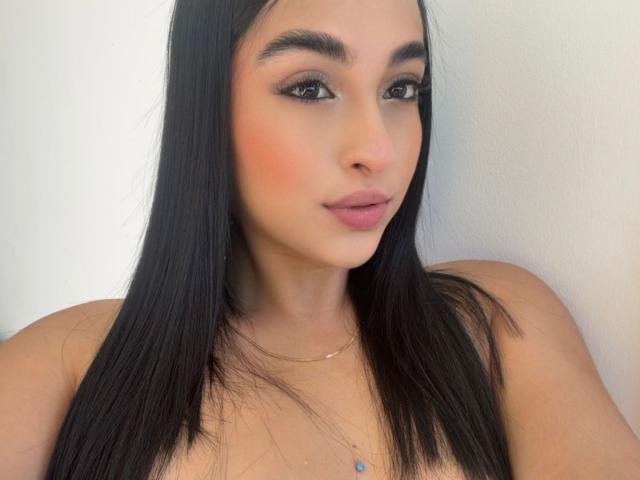 EmmaWaltter with long black hair, brown eyes, pink blush, and pink lipstick, wearing a gold necklace with a blue jewel, showing bare shoulders.