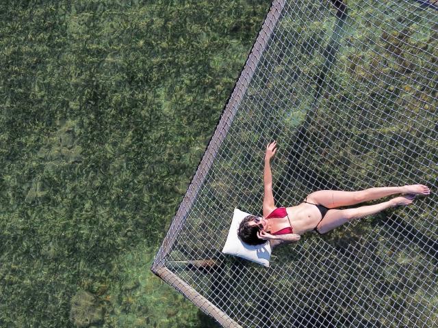 HannaBorbon lying on a white pillow, wearing a red bikini top and black bottoms, with arms and legs stretched out, on a net above clear green water.