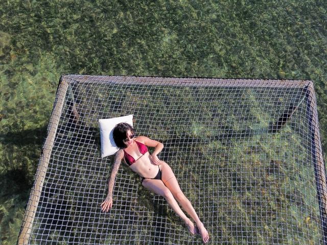 HannaBorbon está flotando sobre una red en agua cristalina, vistiendo un top de bikini rojo y pantalones negros, con un cojín blanco debajo de la cabeza, con los brazos extendidos..
