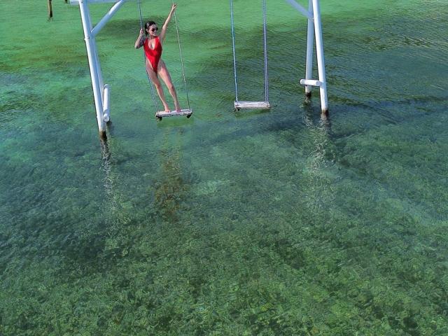 HannaBorbon, con pechos pequeños y un traje de baño entero rojo, está equilibrada en un columpio sobre agua clara y verde, agarrándose al marco de metal del columpio..