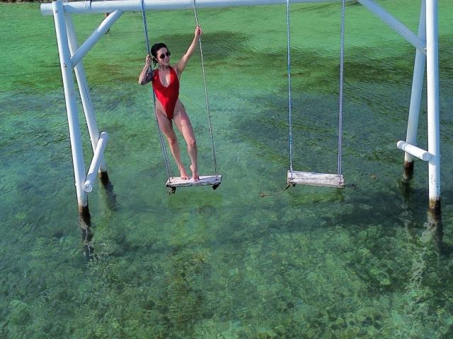 HannaBorbon in a red one-piece swimsuit, standing on a swing in clear green water, tattoo on left arm, holding swing ropes, bare legs, wooden swing, white swing frame.