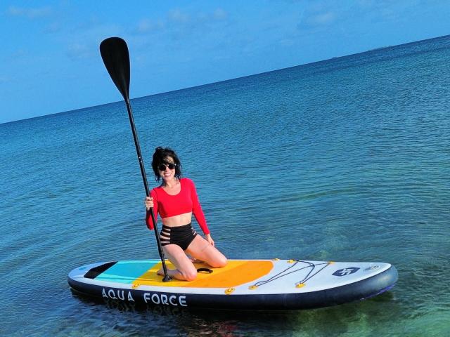 HannaBorbon with tits in a red top, black ass in shorts, kneeling on a paddleboard in clear blue water, holding a black paddle, under a bright blue sky.