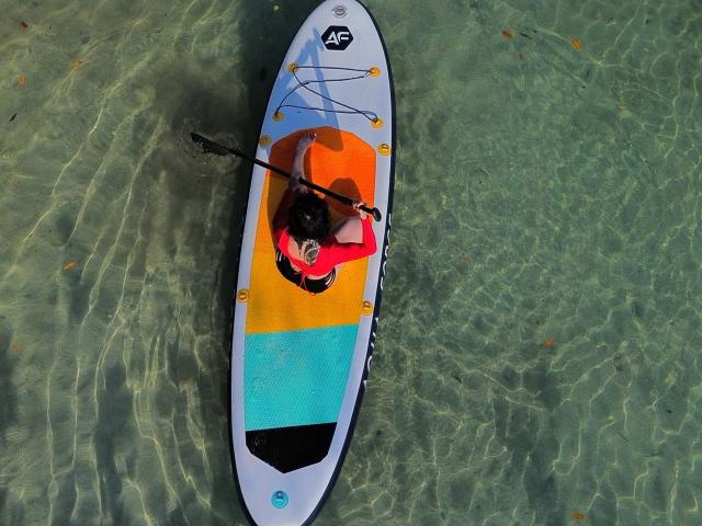 HannaBorbon paddles on a colorful stand-up paddleboard, wearing a red top, black shorts, and a black hat, in clear, rippling water. Paddleboard has yellow, orange, and blue sections.