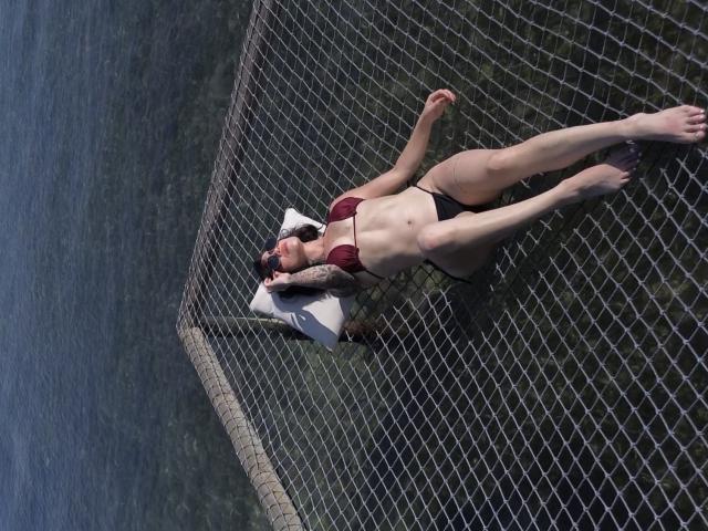 HannaBorbon está acostada sobre una red por encima del agua, vistiendo un top de bikini rojo y pantalones negros, con una almohada blanca debajo de su cabeza. Sus piernas están ligeramente separadas..