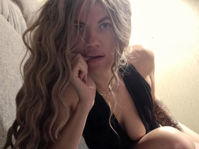 MilaLoona with wavy blonde hair, black dress, and visible tattoos, resting her chin on her hand, showing her tits and a subtle smile, looking at the camera.