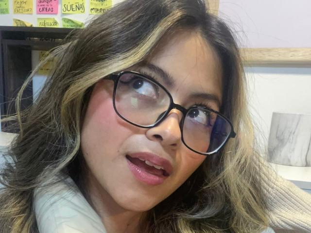 AnittaRouses has medium-length, wavy brown hair with blonde highlights, wearing black glasses, and slightly open pink-lipsticked mouth, showing a bit of her teeth. Her face is pink.