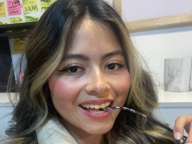 AnittaRouses with wavy blonde and brown hair, smiling, holding a toothbrush in her mouth, pink lipstick, and white shirt, has cute, round ass-like cheeks, medium-sized tits visible.