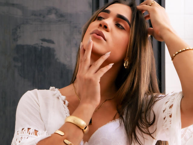 AliceCoolenk with long brown hair, wearing a white lace top, gold earrings, and bracelets, touching her face with manicured nails, looking slightly up with a sultry expression.