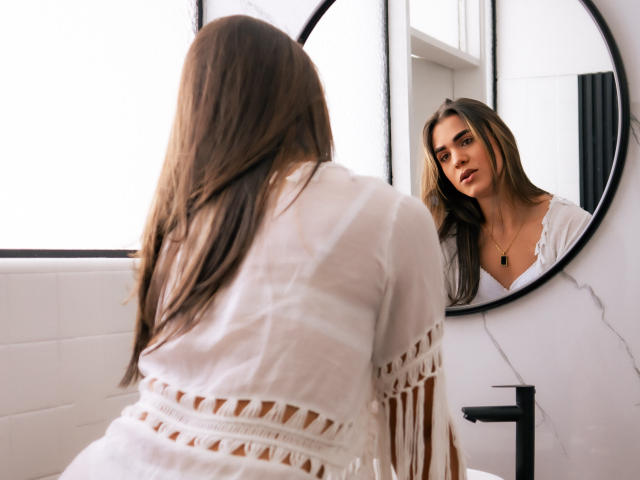 AliceCoolenk with long brown hair, wearing a white fringed top, stands in front of a mirror, reflecting her face and bare shoulders, in a bright, modern bathroom.