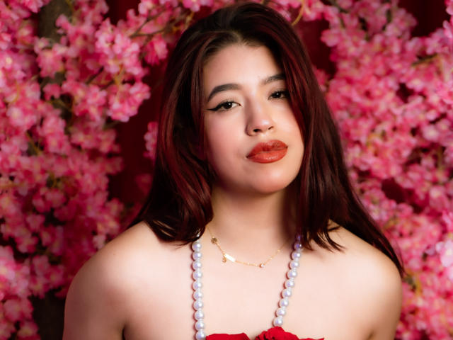 ElizaNior has red lipstick, dark red hair, and is wearing a beaded necklace with white pearls. Her tits are visible, and she's looking at the camera with a slight smile.