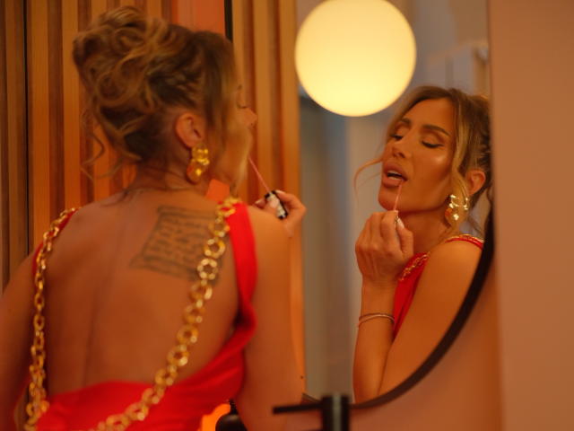 EvelynEvy with wavy blonde hair in a red dress with gold chain straps, applying lipstick in a mirror, showing a tattoo on her back, wearing large gold earrings.