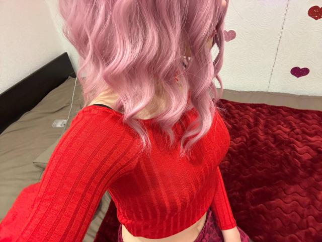 MiyaLia with pink wavy hair wearing a red ribbed crop top, exposing her midriff, sitting on a red quilted bed. Her tits are small, her skin is light.