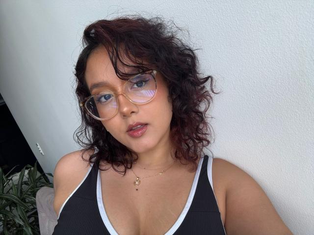 SophiaBrowm with curly dark red hair, wearing glasses and a black tank top with white trim, showing her tits and a gold necklace, standing against a white wall.