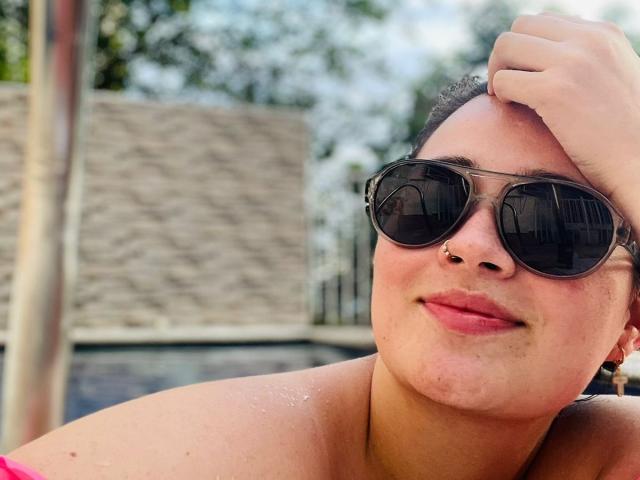 Anny Bentleii, com a pele bronzeada, usando óculos de sol pretos, tem uma fenda charmosa no nariz e um leve sorriso, tocando a cabeça com a mão, mostrando alças rosa nos ombros..