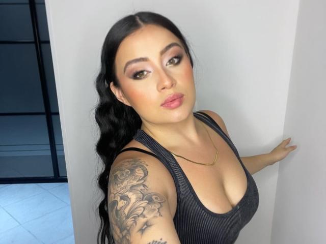 AnnyBentleii mit schwarzen Haaren, braunen Augen und pinkem Lippenstift, trägt ein schwarzes Tanktop, das ihre Brüste zeigt, und hat ein detailliertes Tattoo auf ihrem rechten Arm. Sie steht vor einer weißen Wand..
