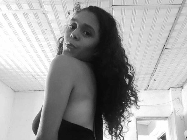 SofiaAndJhonSex with curly dark hair, looking over shoulder, black strapless top, showing her ass, black and white photo, ceiling with grid pattern, soft expression.