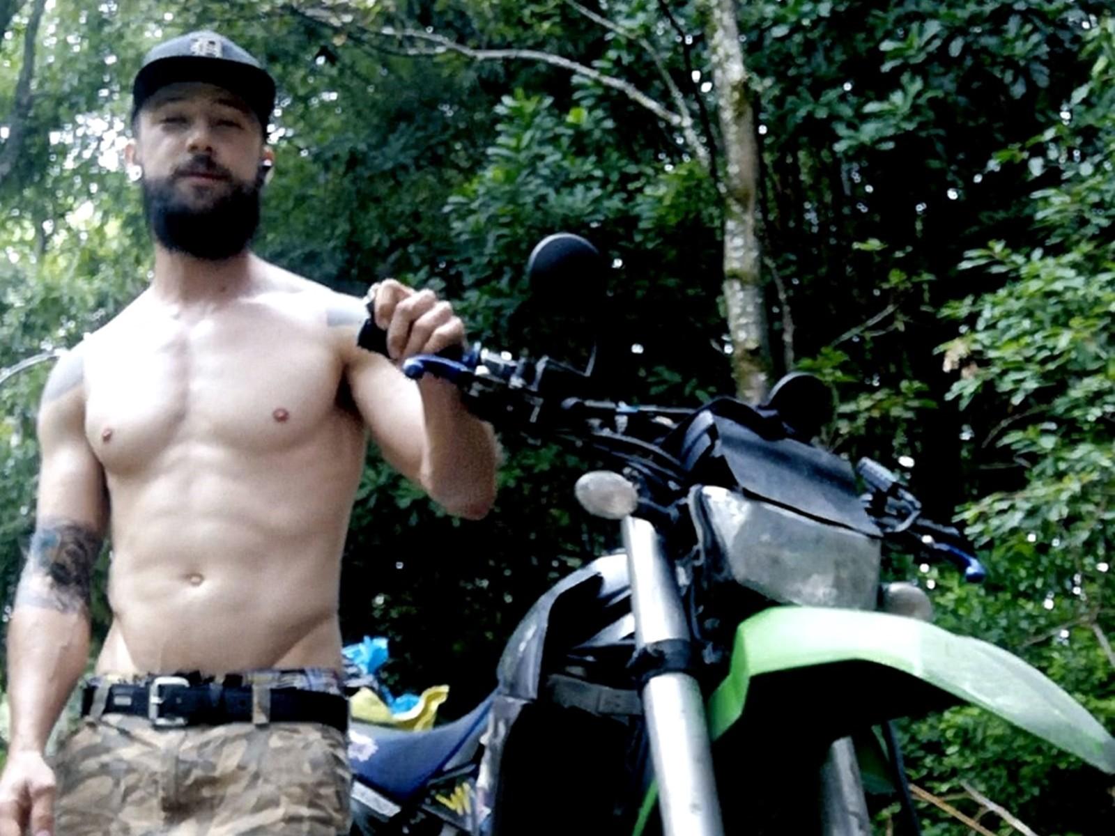 Shirtless LukyXMan with a beard and cap, holding a bike's handlebar, wearing camo pants, and showing a tattoo on his left arm, standing in a forest.